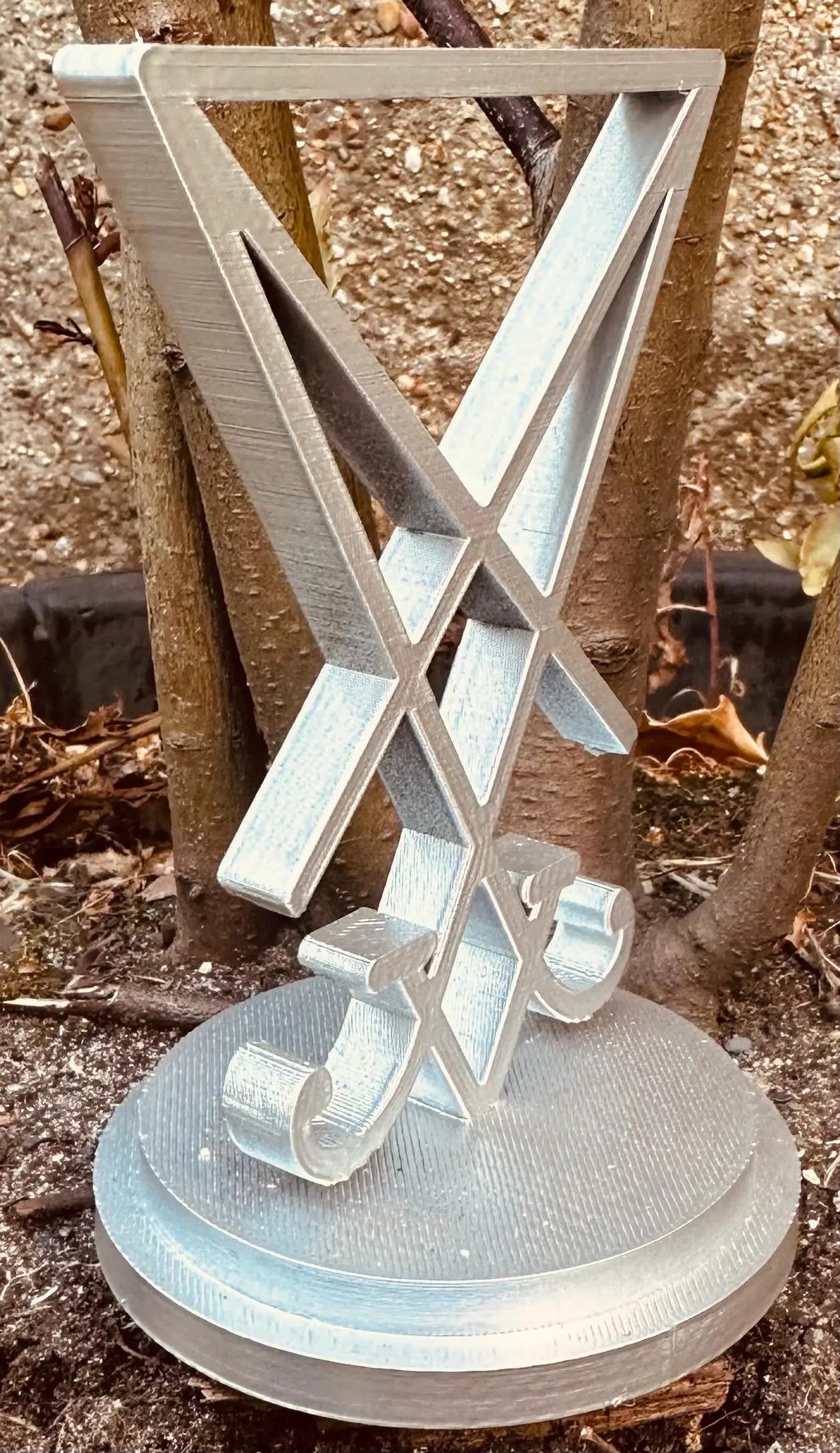 3D printed silver Sigil of Lucifer standing on a round base outdoors against tree bark background