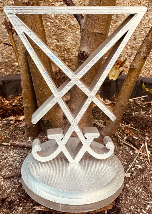 3D printed Sigil of Lucifer in silver on a round base, displayed outdoors against tree trunks and soil background